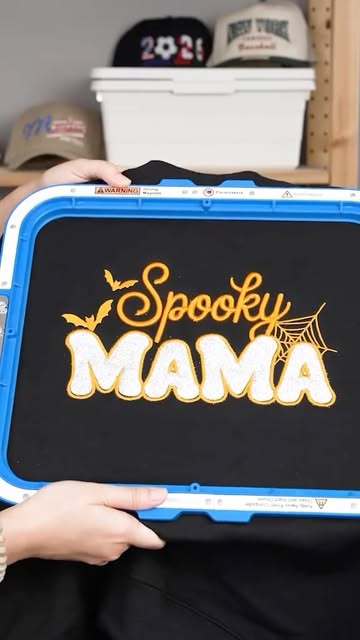 Spooky Mama lettering embroidered on black fabric in a blue embroidery hoop.